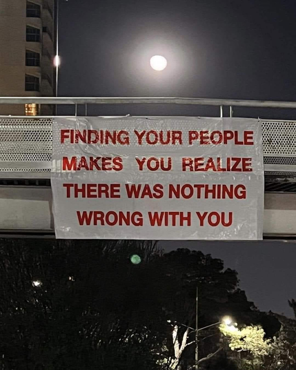 A vinyl sign hanging from an overpass. It says: "Finding your people makes you realize there was nothing wrong with you."