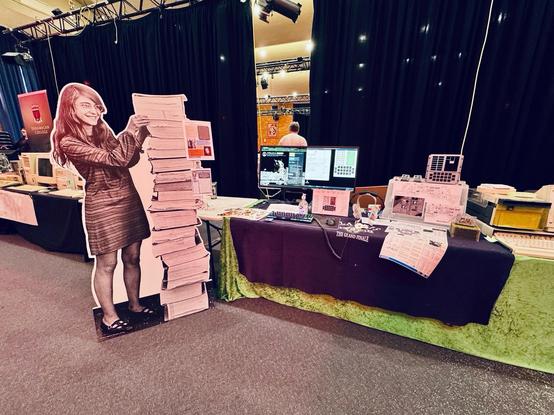 a photo of my complete AGC display, with (L-R) a life-size Margaret Hamilton standee next to the stack of printed-out AGC code, a NASA women of colour poster, a widescreen monitor playing the Apollo in Real Time website and the complete Virtual AGC (inna VM), a life-sized 3D-printed DSKY (WIP, electronics will be in next year) with some cute-as-heck baby mini-DSKYs 😍, and perched atop some magnetic core memory is an accurate replica of the famous SCE to AUX switch 🤓😎