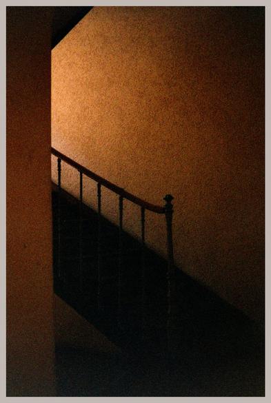 Moody photo of a staircase. Muted colours; sidelit brown wall and a silhouetted railing. Very grainy photo.