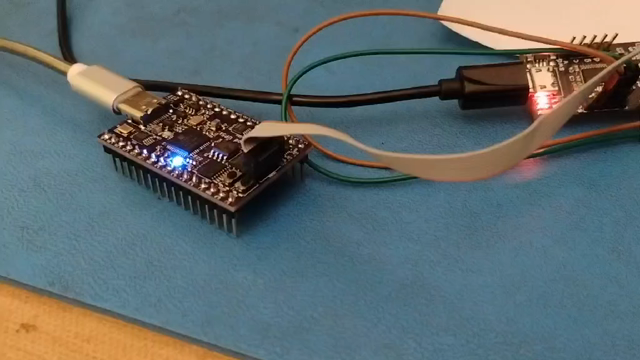 A short video showing two circuit boards laying on a workbench with USB cables heading off to the upper left. Between them is a small gray ribbon cable. The board on the left has a constant blue LED indicating the FPGA is configured, as well as an RGB LED that cycles through seven colors to indicate basic operation.