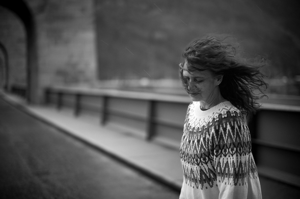 Frau in gemustertem Pullover auf einem Weg vor einer Steinmauer. Starker Wind weht ihr die Haare ins Gesicht, während sie mit geschlossenen Augen nach vorne geht. Schwarz-Weiß-Aufnahme mit ruhiger, nachdenklicher Stimmung.
