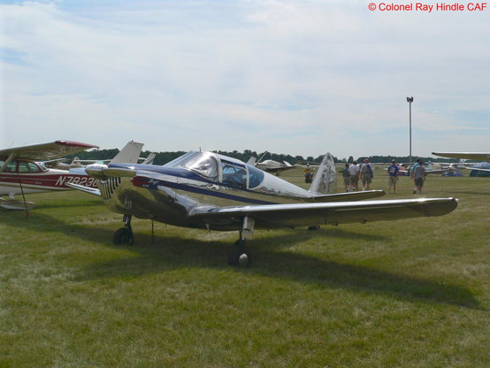Polished Globe/Temco Swift taildragger parked on the grass at Oshkosh 2007. The aircraft has a low metal wing, a framed canopy, and a bright, mirror‑finish fuselage reflecting the surrounding aircraft and visitors. Several other light aircraft and people are visible in the background under a partly cloudy sky