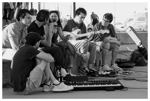 Photo noir et blanc, un groupe de jeunes gens reprennent des chansons, un guitariste et un clavier posé sur le sol