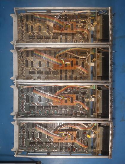 Four electronic devices without their top cover on a workbench seen from above. The circuit boards can be seen, with in each device a small added circuit board with colourful ribbon cables plugged into the original circuit.