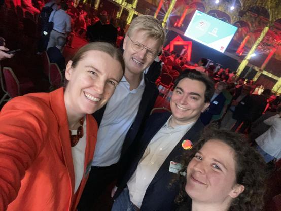 A group of four people poses for a selfie at an event, with a colorful and lively atmosphere in the background. Red lighting and numerous attendees are visible, along with a presentation screen.