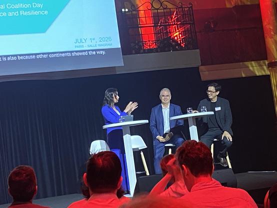 A panel discussion taking place at "National Coalition Day" on July 1, 2026, in Paris. The speaker is gesturing while standing next to two seated panelists, with a screen displaying the event title in the background.