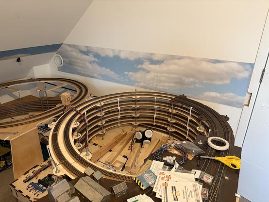 A model train layout in a room, featuring multiple circular tracks and various building materials scattered around. A backdrop with a sky and clouds is visible on the wall.
