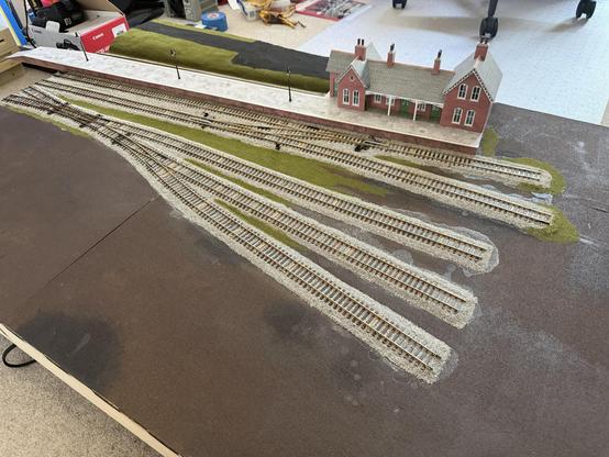 A model railway layout featuring multiple tracks radiating from a central platform. The platform is adjacent to a small station building, with grassy areas and lamp posts. The base is textured to resemble ground, and the scene is set indoors on a tabletop.