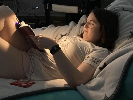 A person is reclining on a bed or couch, reading a book with a small reading light attached. They are wearing a light-colored t-shirt and shorts. A pack of snacks is visible on the surface next to them, and the environment is dimly