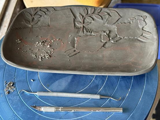 a work in progress carved ceramic tray.  the background is emerging, and two sheep are jumping in the foreground.  An exacto knife and dental tool are visible in the foreground.