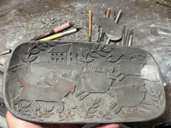 An “almost “ finished carved pottery tray.  You can see a sheep dog chasing 3 sheep in the design.  The top right sheep has a patch over its front feet because I made a dumb mistake.