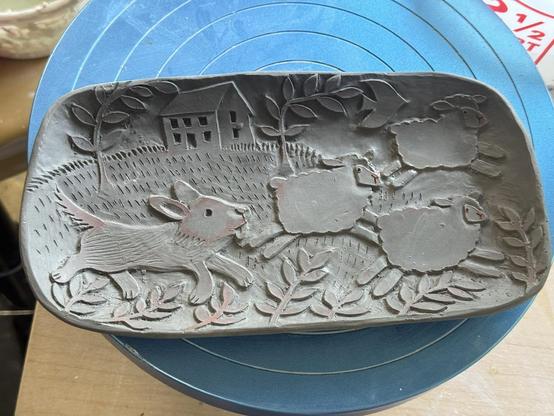 An unfired clay plate. It is decorated with a carving of a dog chasing 3 sheep