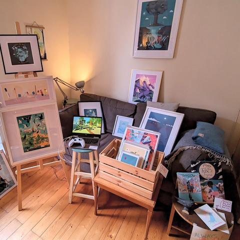 Photo from last year's event showing a living room with a sofa and a easel next to it with framed artworks on both of them as well as the walls. There is a small table with prints in sleeves, another one with books, comics and zines and a bar chair with a small laptop and gamepad running a video game.