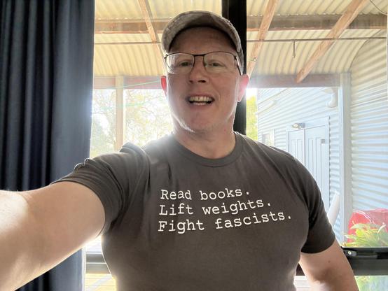 A man in a dark grey tshirt with the words “Read books. Lift weights. Fight fascists.” On the front.