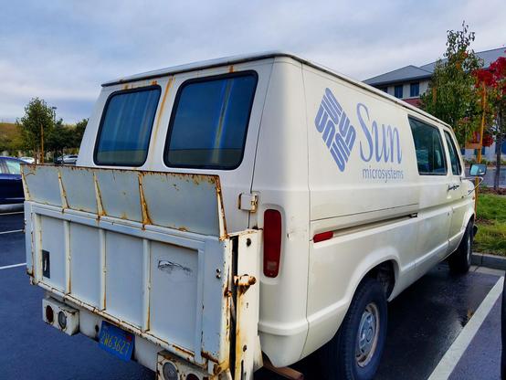 white ford e-series (?) van from the 1970s with a lift up ramp thing on the back and a prominent Sun Microsystems logo on the side, spotted abandoned and rusting in the car park of their former campus