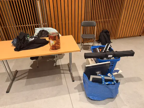 All our gear packed up: An empty 5l beer can on a table, along with some clothes. A large carrying bag with two LCD screens, the Toshiba luggable computer, and a plastic shopping basket with the rest of the parts and gear.
