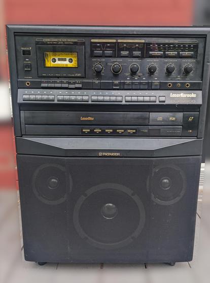 Front showcase of a LaserDisc LaserKaraoke tower, with a big speaker on bottom, LaserDisc player in middle, track selector above, and a Cassette deck with volume control on top.