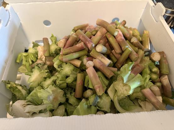 A white wax food service box full of cauliflower stems and leaves, broccoli stems, and asparagus stems.