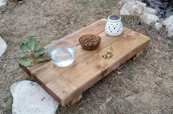 A low table, DIY made with thick wood, on it, a bowl of cat food, a bowl of water, and a solar decorative lamp in white ceramic. Some oak leaves and acorn in decoration.