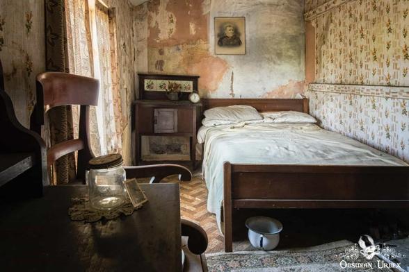 An abandoned bedroom features a wooden bed, vintage nightstand, and distressed wallpaper, with a chamber pot resting under the bed.