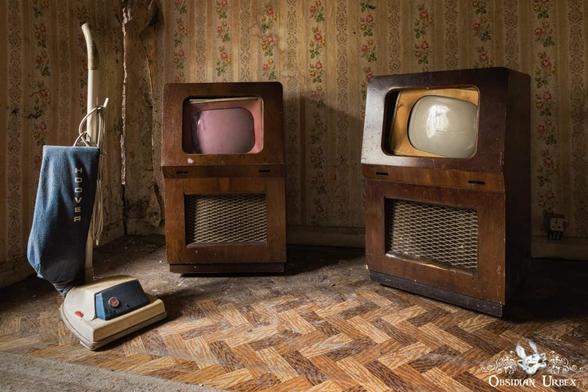 An abandoned room features a vintage Hoover vacuum cleaner and two large wooden console televisions with pink and white screens.