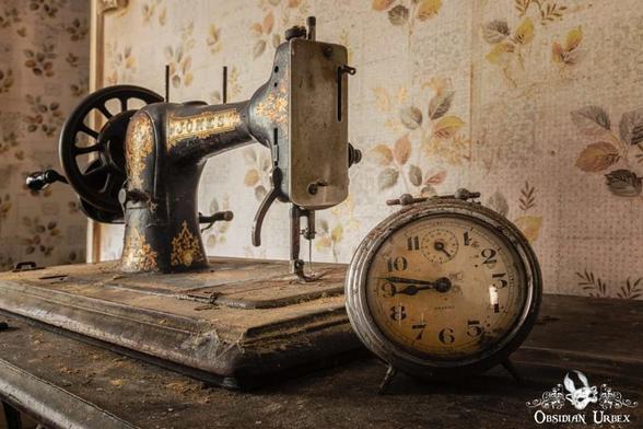 A dusty, vintage sewing machine and alarm clock  rest on a table against floral wallpaper.
