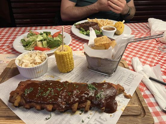 A board on a restaurant table with a big piece of ribs in BBQ sauce, with half of a grilled corn, coleslaw and potato chips. The other person has a steak with fries, corn and salad.
