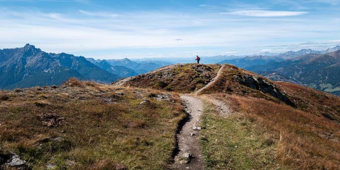 Blick von einer Bergspitze. Ein schmaller Schotterweg führt auf eine noch mal etwas höhere Anhöhe. Darauf ein Wanderer mit Wanderstöcken und roter Jacke. Im Hintergrund von linker bis rechter Bildseite Alpenpanorama mit teils schroffen Berggipfeln oberhalb der Baumgrenze und Bäumen weiter unten an den Bergen. Weitgehend blauer Himmel mit dünner, fast gemalter Wolkenschicht.