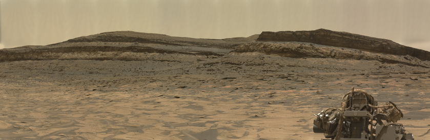 An ochre toned image capturing a Martian landscape, with the instrumented turret of the Mars rover Curiosity in the foreground.