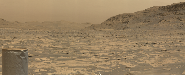 An ochre toned image capturing a Martian landscape, with the UHF antenna of the Mars rover Curiosity in the foreground.