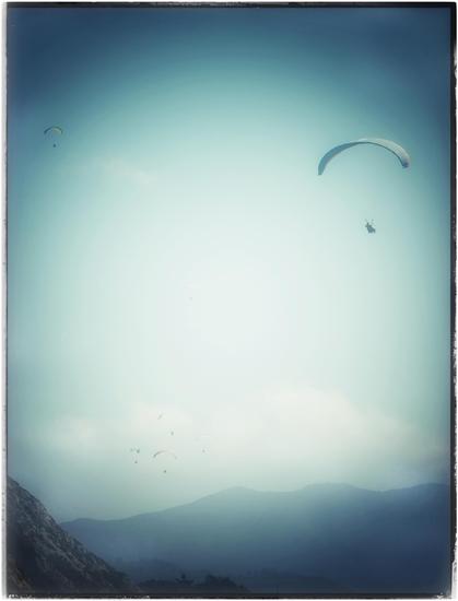 A misty, teal-tinted view of half a dozen paragliders floating through the sky, above the coastal mountains and an unseen ocean below. It's a peaceful scene of mostly empty sky and a few small, faded mountains. Taken from the cliffs of Thornton State Beach, just south of San Francisco, where the wind hits the cliff edges and rises upward on a daily basis.