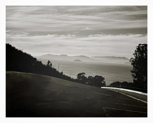 A serene black and white landscape photograph of a hill overlooking a vast bay with islands in the distance and vibrant wispy clouds floating above. We are looking out at infamous Alcatraz Island and the San Francisco Bay from high atop Yerba Buena Island. In the distance behind you can see Sausalito and the Marin Headlands, Angel Island, and just the tiniest hint of the Golden Gate Bridge. In the foreground is a very sloped, abandoned parking lot bordered by two roads heading down the hill, converging at the bottom and providing a nice forced-perspective leading the eye to that tiny little Alcatraz floating out there is the fog.
