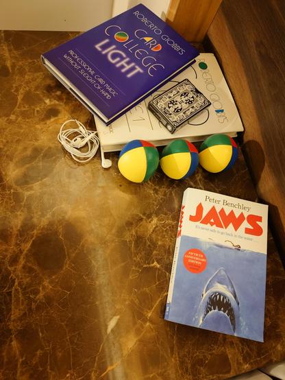A photo of a brown side table next to a bed. First is the book version of Jaws, which is a bold choice for a seaside destination. Above that is 3 juggling balls, some wired earphones, a deck of cards and 2 books about learning card magic.