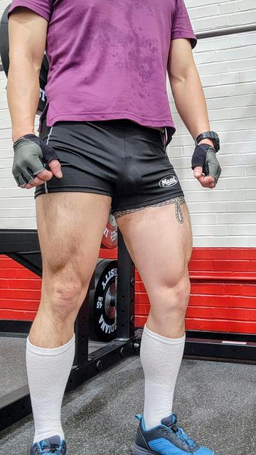 Legs standing in front of a squat rack. Short black shorts, long white socks, purple t-shirt, gray fingerless gloves, blue sneakers, cage print.