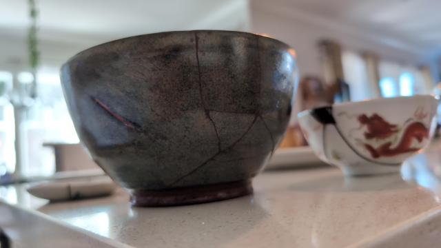 A large bowl with dark cracks tracing its surface. Behind it is a smaller bowl with a dragon motif, with a dark chip along the lip and two large cracks leading away from it.