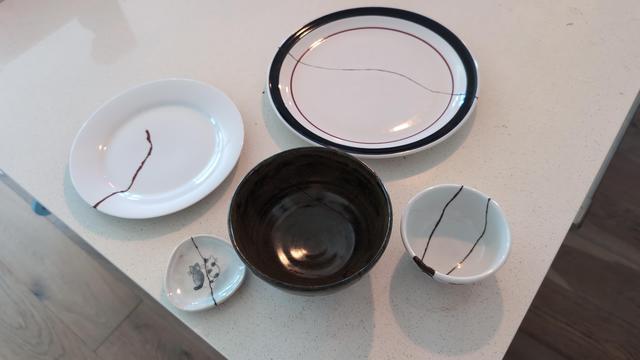 A photo from up higher. The two bowls in the previous photo are visible, along with three plates. The plates all have cracks down their middles. The smallest has two cats sitting together painted on it; the crack runs right between them.