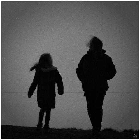 Sur fond de ciel sombre, deux silhouettes marchent vers nous, une femme et sa jeune fille. Elles ont le visage tourné l'une vers l'autre et semblent discuter.