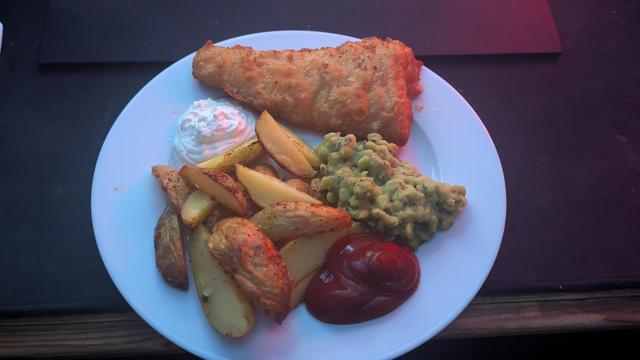 A white dinner plate on a black background. On the plate is a large piece of beer batter cod, some minty mushy peas and some crispy home made seasoned wedges made from salad potatoes. On either side of the plate is a dollop of mayo and ketchup.