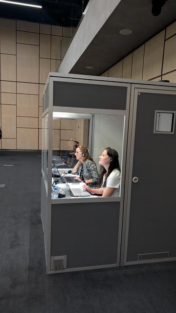 Two translators in a small container with a glass front at the back of the conference room. They have a mic and a laptop in front of them looking at the stage translating live.