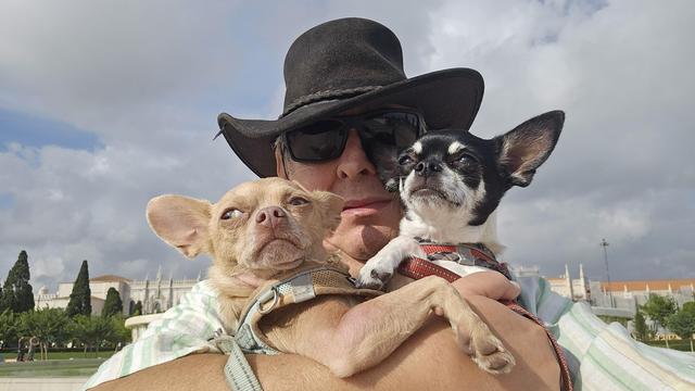 A man wearing a dark hat and sunglasses holds two small Chihuahuas in a selfie. The tan Chihuahua on the left looks up and to the right with a slightly worried expression, while the black and white Chihuahua on the right looks up with a more serious gaze. In the background, under a partly cloudy sky, a large white building with intricate architecture and green trees is visible.