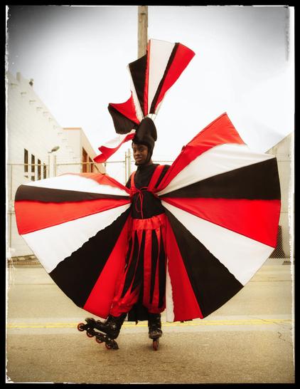 A photograph of a teenaged boy on rollerblades. He is dressed for a street fair, wearing a bright red, black, and white costume with a fan-like cape and matching, tall, plumed hat. He paused briefly for the camera, facing me and spreading his cape, wings to the breeze, before skating off to join the celebration.
