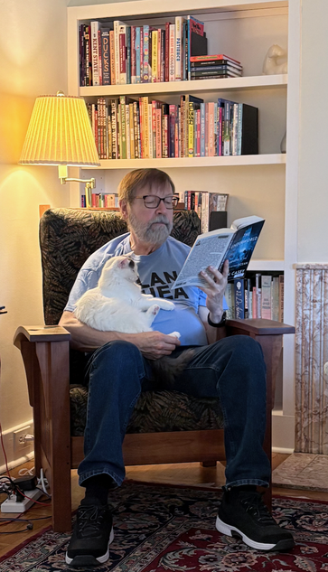 A man sitting in a chair reading a book that he's holding in one hand. the other arm is wrapped around a white cat that's sitting on his lap.