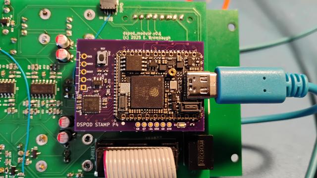 A color photograph of a small purple PCB mounted on a larger green PCB. The purple PCB has a smaller black PCB mounted on it via castellated pads and a blue USB-C cable plugged into the right side. At the bottom of the image a gray ribbon cable is seen extending out of frame.