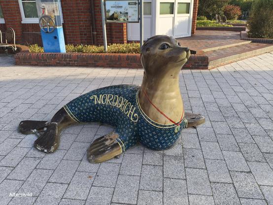 Skulptur am Touri-Infocenter in Norddeich: eine Robbe in einem Blauen Kleid (?) mit gelbem Floramuster. Sie trägt ein rotes Band mit einem Anhänger, auf dem das N-Logo der Stadt Norden zu sehen ist. Auf dem Kleid steht: Norddeich