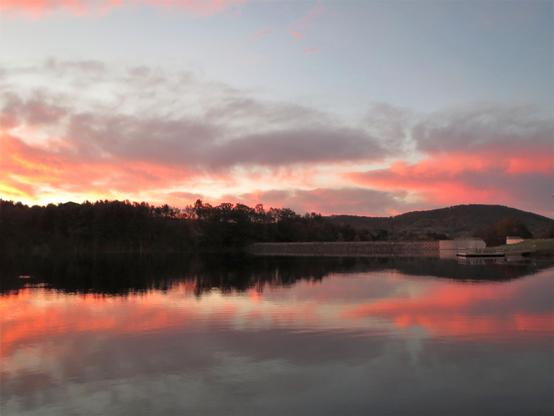 The lake we have so often visited and photographed is manmade. I rarely take photos of the dam area, but on this particular morning the sunrise clouds suggested I do so. The spillway is to the extreme right and the stone and earthen dam extends out to the left toward a line of trees. In the background is the head of a ridge, a hill known as Donats Peak. All described is reflected on the surface of the lake.