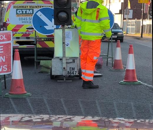 A temporary signal in the road, with cones around and a man in hi-vis stationed nearby. The signal box has serial number EM 8647.