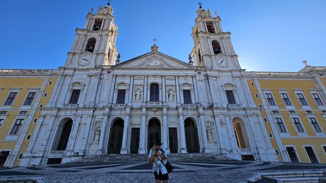 A low-angle, wide shot captures the grand, white Baroque facade of the Mafra National Palace under a clear blue sky. Two prominent bell towers with clocks flank the central church entrance, which features statues in niches and ornate details. A person wearing a hat and holding a small dog stands on the patterned cobblestone plaza in front of the palace, which also has long yellow wings extending from the central structure.