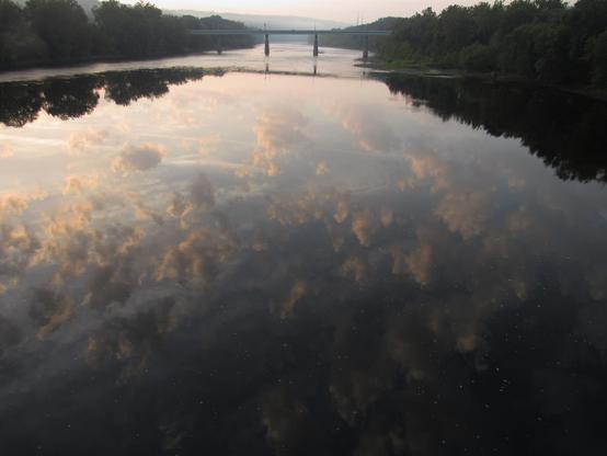 Cloud reflections from the largely unseen sky above appear on the surface of waters of the Delaware river which creates a border between New Jersey and Pennsylvania. Trees line the shores on either side. There is a bridge in the distance.