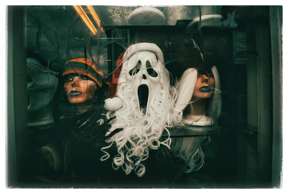 A view through a graffiti-etched, green-tinted window pane, looking at three mannequin heads on a glass shelf. Two of the heads belong to a pair of icy women who are staring coldly out onto the sidewalk. The middle head, however, belongs to Santa Claus. Santa has a traditional red cap and white curly beard... and an unconventional mask from the movie Scream. The mask makes Santa look like he is howling into the void.

Aren't we all?