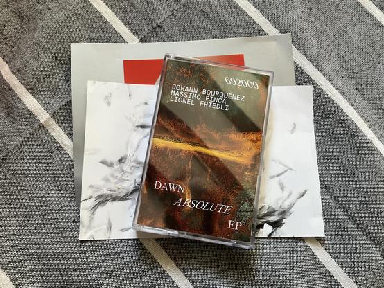 Cassette du nouvel EP de Johann Bourquenez, posée sur des cartes postales sur un tissu gris à rayures blanches.
